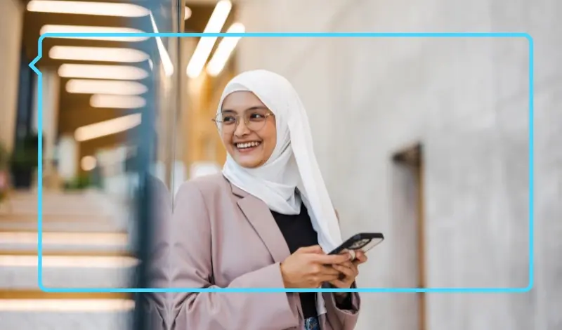 Smiling woman in a white hijab and pink blazer holding a smartphone while standing on modern indoor steps, framed by a blue speech bubble